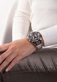 Srebrn ton ure s velikim okroglim številčnikom, jasnimi merilnimi površinami, dodatki, podobnimi diamantom, in kovinskim povezovalnim zapestjem. Nošena na roki, ki počiva na teksturirani površini.