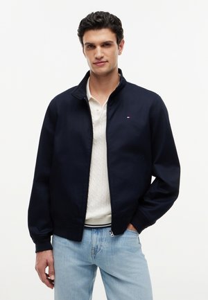 Mężczyzna o ciemnych włosach ubrany w granatową kurtkę na zamek błyskawiczny na białym dzianym swetrze i jasnoniebieskich dżinsach, stojący z jedną ręką w kieszeni.