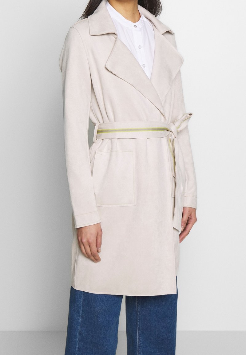 Ljusbeige faux suede trenchcoat med bälte i midjan, slagkrage och sidofickor. Har en slät yta och rak skärning.