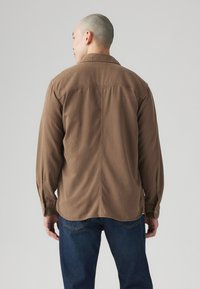 Camisa de algodón marrón con mangas largas, cuello con solapa y puños abotonados. Espalda plana con una textura suave y detalles de costura sutiles.
