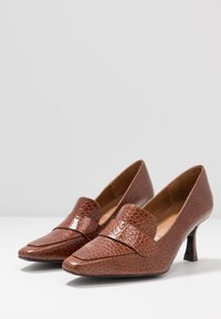 Tacones de cuero marrón con un acabado texturizado, punta afilada y una banda ancha en la parte frontal. Tacón alto y delgado con una superficie lisa.