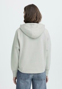 Felpa grigia chiara in cotone con cappuccio regolabile, spalle scese e polsini a coste. Abbinata a jeans blu a vestibilità morbida.
