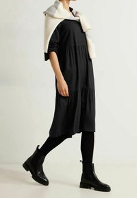 Schwarzes, gestuftes Kleid aus weichem Material mit langen Ärmeln und seitlichen Taschen. Ein creme- und graufarbenes Strickpullover, der über die Schultern drapiert ist.