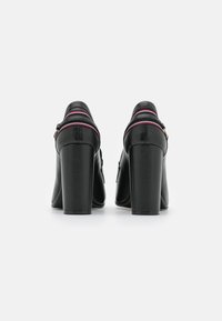 Zapatos de tacón alto de cuero negro con un tacón grueso, con costuras visibles y acentos de colores en la correa. Textura suave, vista trasera.