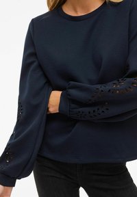 Maglione a maniche lunghe blu navy con ricami a fiori intagliati sulle maniche a sbuffo, indossato con pantaloni neri e le braccia incrociate in vita.