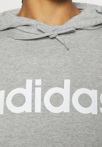 Sweat à capuche gris avec un grand logo blanc "adidas". Le matériau est doux avec une finition texturée. Comprend un cordon de serrage au niveau de la capuche.