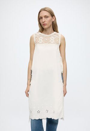 Femme portant une robe blanche sans manches avec un panneau supérieur en dentelle et un ourlet découpé floral, associée à un jean bleu, debout devant un fond uni.
