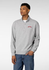 Grå hettejakke med glidelås laget av mykt stoff, med h�øy krage og rød brodert "HENRY CHOICE"-logo på brystet.