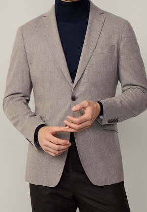 Blazer ajusté gris clair en tissu texturé, fermeture à un bouton, revers cranté, et poche plaquée, porté sur un col roulé bleu marine.