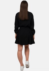 Abito nero con maniche lunghe e gonna svasata; tessuto texturizzato, cintura liscia, abbinato a sneakers bianche. Vista posteriore mostrata.