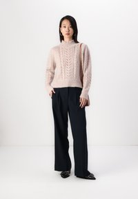 Csipkés kötésű, rózsaszín pulóver magas gallérral, sötétkék, bő szárú nadrággal és fekete, hegyes orrú cipővel párosítva. A modell egy barna táskát tart.