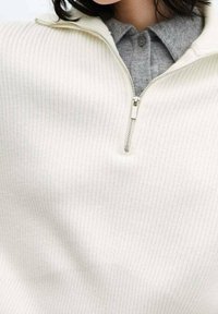 Pull en maille blanche avec un col zippé mi-haut. En dessous, une chemise à col grise est visible. Le tissu semble doux et texturé.