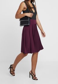 Una gonna bordeaux profondo scende da una vita aderente, abbinata a una borsa a tracolla con perline nera. Tacchi a spillo neri completano il look.