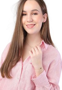 Une jeune femme porte une chemise à rayures rose clair, un collier délicat en or avec des perles, et un bracelet en or avec un petit pendentif.