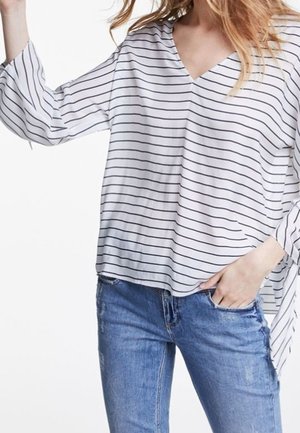 Kvinde iført en hvid, langærmet V-halsbluse med tynde sorte vandrette striber og blå jeans, med den ene hånd i lommen, armen let løftet.