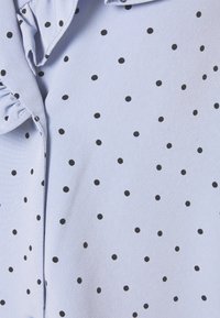 Tessuto azzurro chiaro con piccoli pois neri distribuiti uniformemente e una cucitura del colletto parzialmente visibile sul lato sinistro.