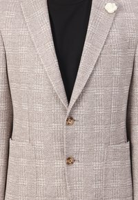 Beige strukturierter Blazer mit einem dezenten Karomuster, ausgestattet mit zwei Fronttaschen und braunen Knöpfen. Inklusive einer weißen Blumen-Anstecknadel.