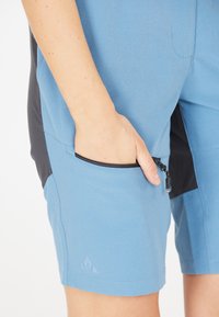 Siniset shortsit, joissa on sileä pinta, musta paneeli ja vetoketjullinen tasku. Sisältää kolmion muotoisen logon helman lähellä.