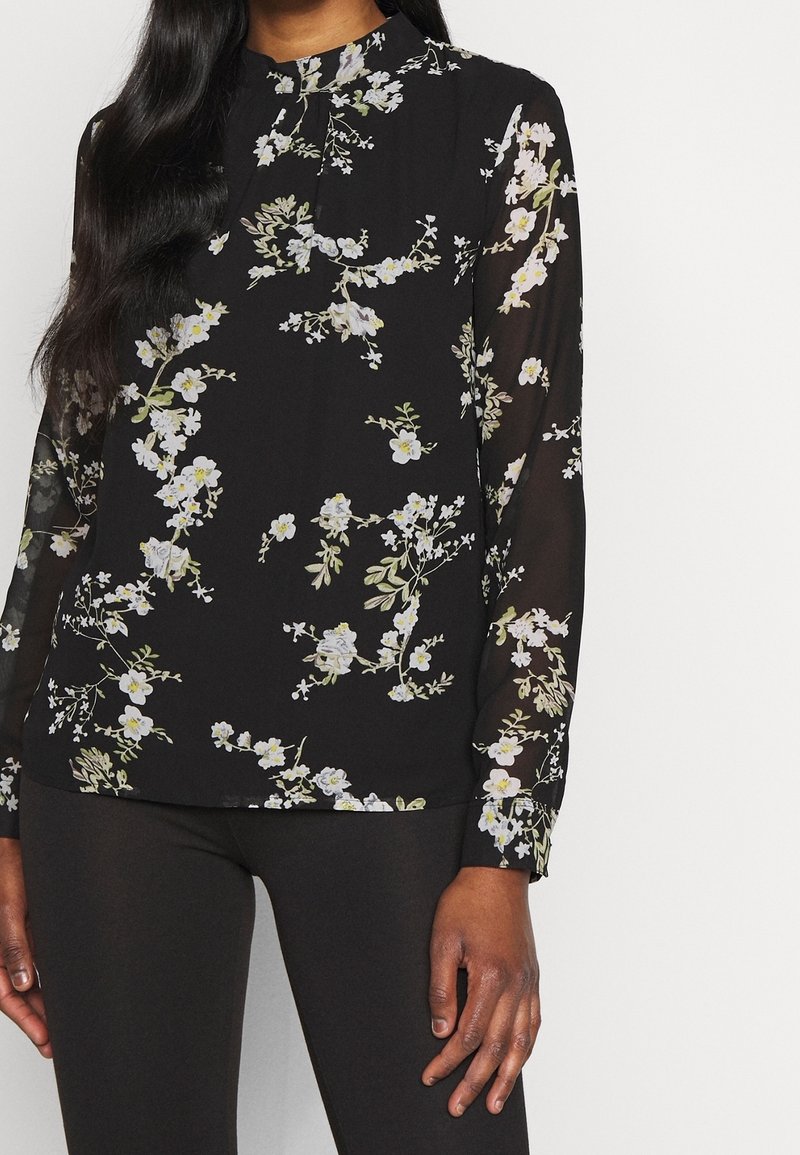 Femme portant un chemisier noir transparent à manches longues avec imprimé floral blanc et col montant, associé à un pantalon noir ajusté.