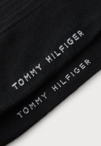 Chaussettes noires avec du texte tissé blanc qui dit "TOMMY HILFIGER." Texture lisse avec un design standard, de longueur cheville.