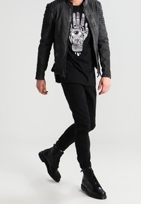 Czarna skórzana kurtka z żebrowanymi detalami na ramionach, w połączeniu z czarnym t-shirtem z grafiką w białym kolorze, dopasowanymi czarnymi spodniami oraz czarnymi botkami.