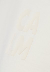 Weißer Stoff mit glatter Textur zeigt erhabene, cremefarbene Buchstaben, die "CALM" schreiben. Das Design ist minimalistisch und modern.