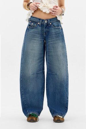 Mujer con vaqueros azul de pierna ancha y dobladillos recogidos sobre zapatos de senderismo marrones, levantando un top blanco corto para mostrar el abdomen.