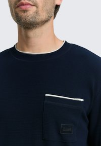 Dunkelblaues Sweatshirt mit Rundhalsausschnitt aus strukturiertem Stoff, ausgestattet mit einer weißen, umrandeten Brusttasche und einem kleinen schwarzen Logotag.