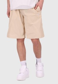 Shorts beiges avec un motif texturé, poche latérale et petit logo blanc. Portés avec des baskets blanches et des chaussettes à motifs.
