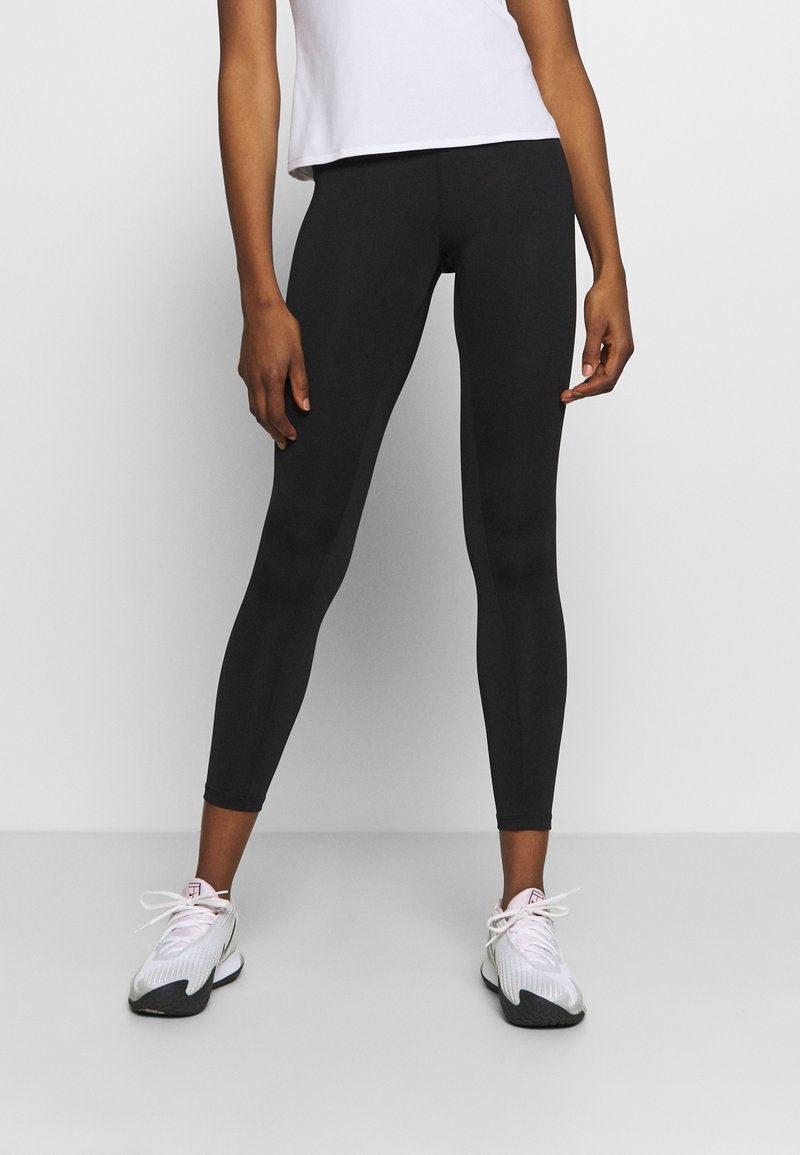 Schwarze Leggings aus dehnbarem Material, eng anliegendes Design, glatte Textur, kombiniert mit weißen Sneakern mit gemustertem Obermaterial und Schnürsenkeln.