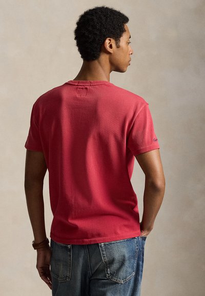 Jeune homme aux cheveux courts et bouclés portant une chemise rouge à manches courtes et un jean bleu, debout de dos sur un fond uni.