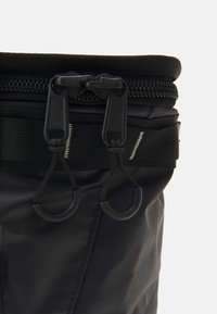 Black waterproof bag with a textured finish, top zipper closure, and two pull tabs. Features a sturdy strap and visible stitching details.