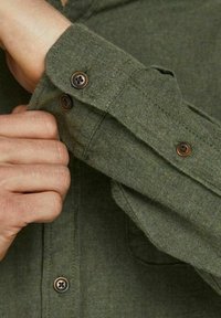 Person som knäpper knappen på ärmen av en grön långärmad skjorta med synlig sömnad och bruna knappar.