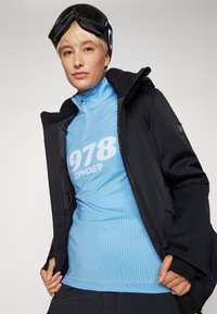 Giacca nera isolata sopra un maglione azzurro chiaro strutturato con mezza zip e grafica bianca. Modello con berretto nero e occhiali protettivi.