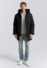 Parka nera isolata con cappuccio, dotata di cerniera frontale, tasche applicate e vestibilità comoda. Indossata sopra una maglia grigia e jeans chiari.