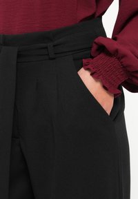 Black trousers featuring a cinched waistband with a belt, side pockets, and a smooth texture. The top has a ruffled burgundy sleeve.