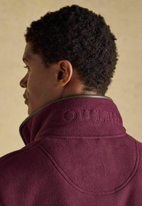 Bordová fleecová bunda s vysokým golierom, na zadnej strane so vzorovaným nápisom "JOULES" a jemnými kontrastnými detailmi.