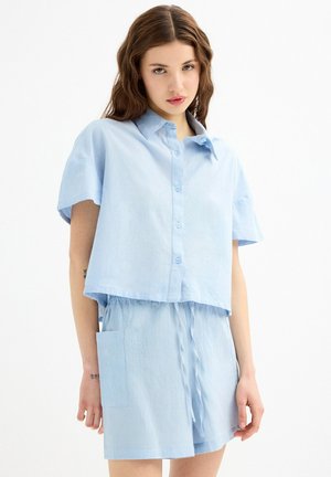 Camisa corta de color azul claro con mangas cortas y cierre de botones. Corte holgado con un bolsillo lateral y tejido texturizado.