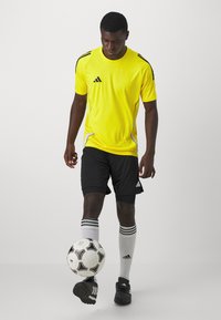 Maillot de sport jaune avec accents noirs, shorts noirs, chaussettes de soccer blanches avec rayures, et baskets noires. Ballon de soccer aux pieds.