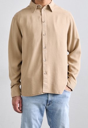 Homme portant une chemise beige à manches longues boutonnée et un jean bleu clair, une main dans la poche et l'autre pendant.