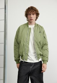 Giacca bomber verde oliva in materiale leggero, con chiusura frontale a zip, polsini a coste e un piccolo logo nero sul petto.