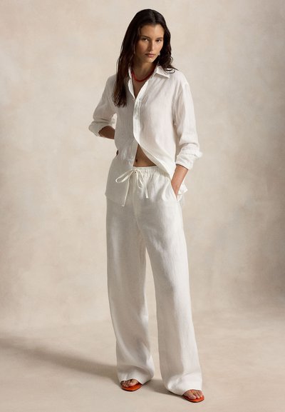 Γυναίκα με χαλαρό λευκό λινό πουκάμισο και παντελόνι, φορώντας πορτοκαλί σανδάλια, στέκεται με το ένα χέρι στην τσέπη μπροστά σε ουδέτερο φόντο.