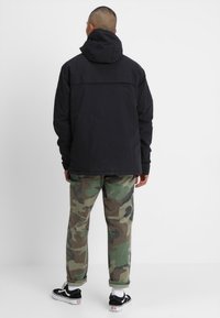 Man som står med ryggen vänd, iklädd en svart huoodad jacka, camouflagemönstrade byxor, vita strumpor och svarta Vans sneakers på en vit bakgrund.
