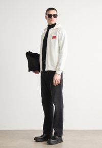 Felpa bianca con zip e logo rosso, dolcevita nero, pantaloni neri e scarpe nere voluminose; modello che tiene una borsa per indumenti nera.