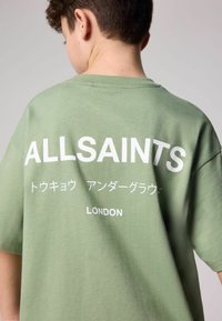 Zaļš kokvilnas T-krekls ar īsām piedurknēm. Uz muguras balti izdrukāts "ALLSAINTS" un citi teksti. Vienkāršs, brīvs dizains.