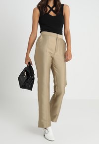 Pantaloni beige su misura con vita alta, tasche frontali e dettagli di cucitura, abbinati a un top nero senza maniche e stivali bianchi alla caviglia.
