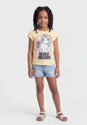 Fille aux cheveux tressés portant un t-shirt jaune avec une licorne colorée et le texte "Beach Dreams", un short en jean et des sandales blanches, debout.