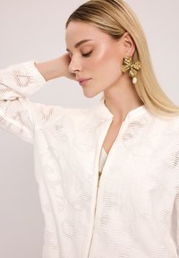 Biała kurtka o fakturze z zamkiem z przodu, z delikatnym wzorem. Duże złote kolczyki w kształcie kwiatu z perłowym akcentem uzupełniają stylizację.