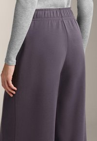 Fialové kalhoty s širokými nohavicemi a strukturovaným povrchem, elastickým pasem a bočními švy. Spárovány s světlým šedým dlouhým tričkem s proužky.