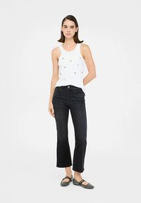 Top blanco sin mangas con pequeños detalles metálicos, combinado con vaqueros acampanados azul oscuro con sutiles adornos en la parte delantera.